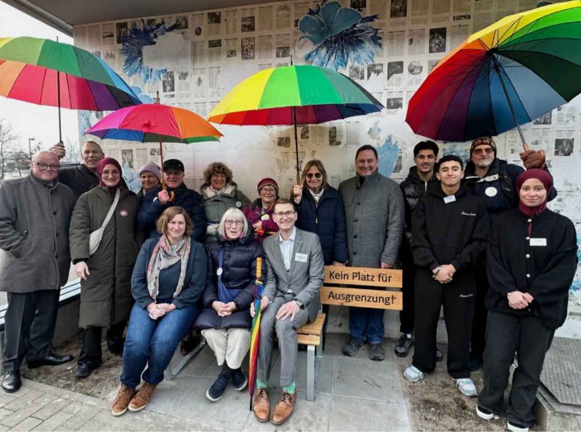 Unter einem überdachten Bereich auf dem Schulhof steht eine gemischte Gruppe aus Erwachsenen und Jugendlichen dicht zusammen. Zwei Personen sitzen vorne, daneben ist eine Holzbank mit Schild zu sehen: ‚Kein Platz für Ausgrenzung!‘ Mehrere Personen halten große, bunte Regenbogen-Schirme als Symbol für Vielfalt. Im Hintergrund befindet sich eine Wand mit Collage/Plakatgestaltung. Das Foto dokumentiert die feierliche Einweihung der Bank als Zeichen gegen Ausgrenzung und Diskriminierung.