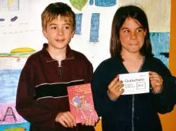 Zwei Kinder halten ein Buch und einen Gutschein in die Kamera, dahinter eine bemalte Wand.