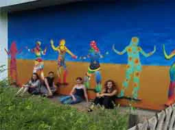 Ein großes Wandbild mit tanzenden Figuren auf blauem und orangem Grund, davor sitzen die Gestalterinnen und Gestalter.
