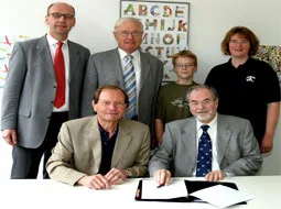 Bei der Vertragsunterzeichnung sitzen zwei Männer am Tisch, dahinter stehen Kinder mit Blasinstrumenten.