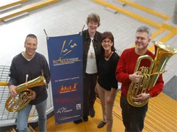 Zwei Musiker halten Tubas, daneben stehen zwei Frauen vor einem Banner der Bläserklasse.
