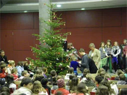 In der Aula steht ein großer Weihnachtsbaum, darum herum sitzt und steht das Publikum.