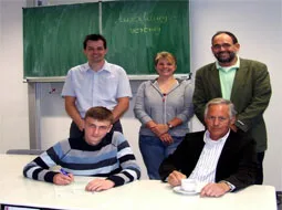 Ein Schüler unterschreibt am Tisch seinen Ausbildungsvertrag, daneben stehen die Beteiligten.