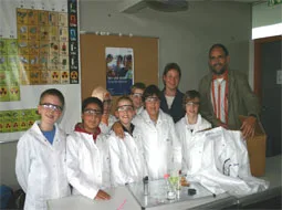 Kinder in weißen Laborkitteln stehen mit Lehrkräften am Experimentiertisch.
