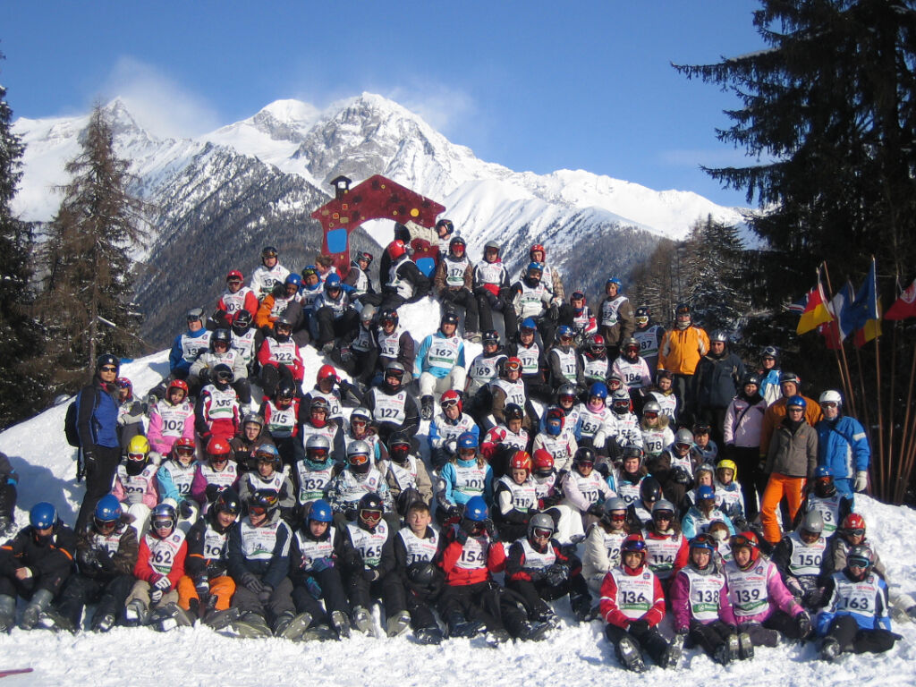 Eine große Gruppe in Skikleidung mit Startnummern sitzt und steht auf einem verschneiten Hang; im Hintergrund schneebedeckte Berggipfel und Flaggen.