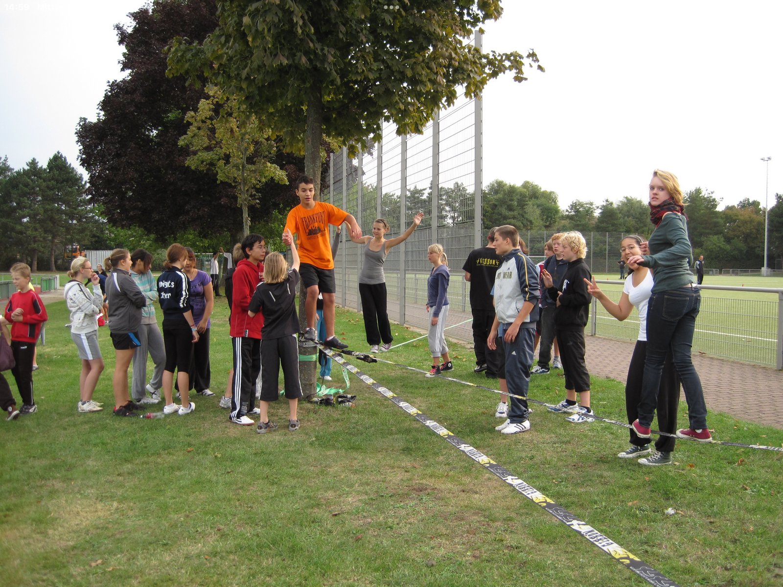 Jugendliche balancieren auf einer zwischen einem Baum und einem Pfosten gespannten Slackline auf einem Sportplatz; eine Gruppe schaut zu.