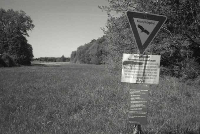 Ein Naturschutzgebiet-Schild „Kammereckswiesen bei Langen“ am Rand einer von Bäumen gesäumten Wiese (Schwarz-Weiß-Aufnahme).
