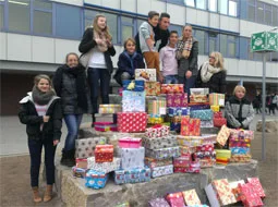 Jugendliche stehen hinter einem großen Stapel bunt verpackter Päckchen vor dem Schulgebäude.
