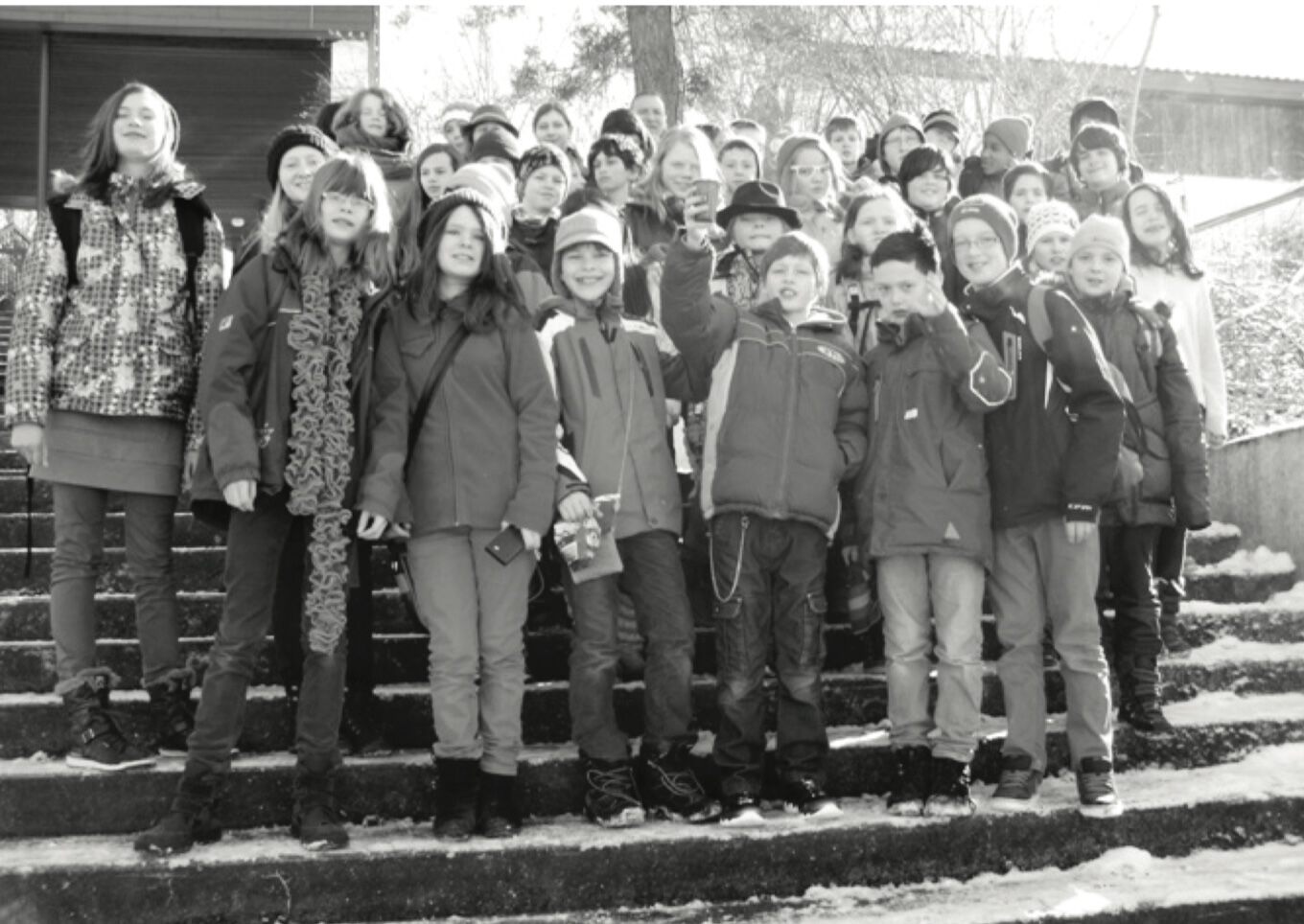 Eine große Gruppe Kinder in Winterkleidung steht auf verschneiten Außentreppen; einige lächeln in die Kamera (Schwarz-Weiß-Aufnahme).