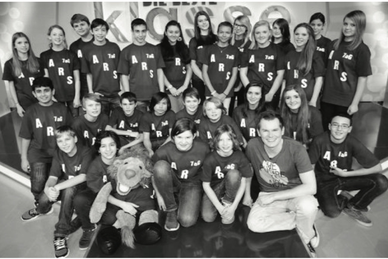 Eine Schulklasse in einheitlichen „ARS 7aG“-T-Shirts posiert in einem Fernsehstudio mit einem Maskottchen und dem Moderator vor der Kulisse „Die beste Klasse Deutschlands“ (Schwarz-Weiß-Aufnahme).