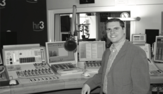 Eine Person im Anzug steht lächelnd in einem Radiostudio des Senders hr3 vor Mischpulten, Bildschirmen und einem Mikrofon (Schwarz-Weiß-Aufnahme).