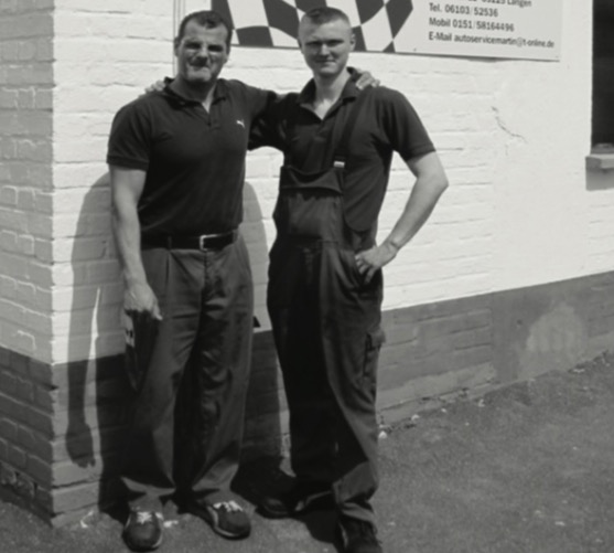 Zwei Männer stehen Arm in Arm vor einer Kfz-Werkstatt; einer trägt eine Arbeitslatzhose, an der Wand ein Schild mit Zielflaggen-Logo (Schwarz-Weiß-Aufnahme).