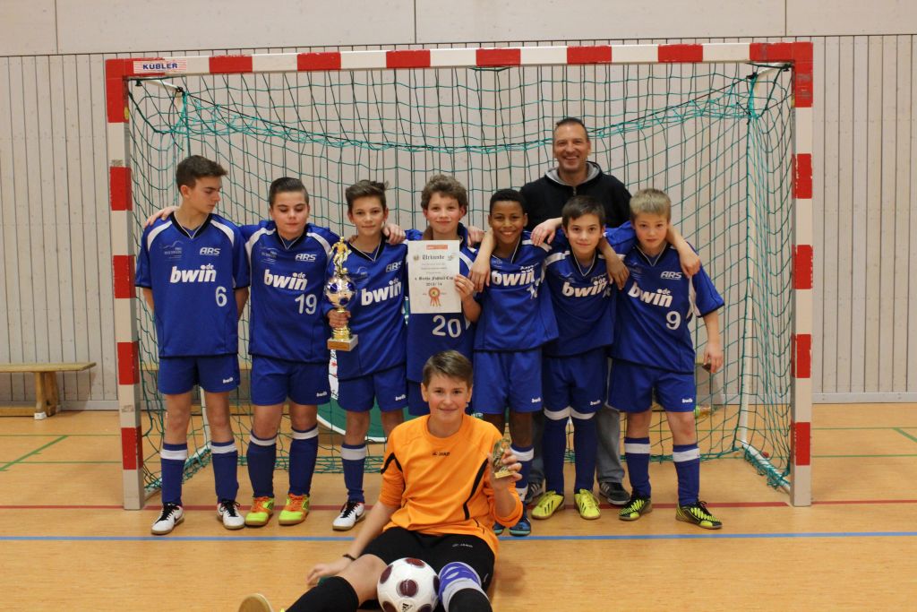 Eine Fußballmannschaft in blauen ARS-Trikots posiert mit ihrem Trainer vor einem Hallentor; sie halten einen Pokal und eine Urkunde, vorne sitzt der Torwart in Orange mit dem Ball.