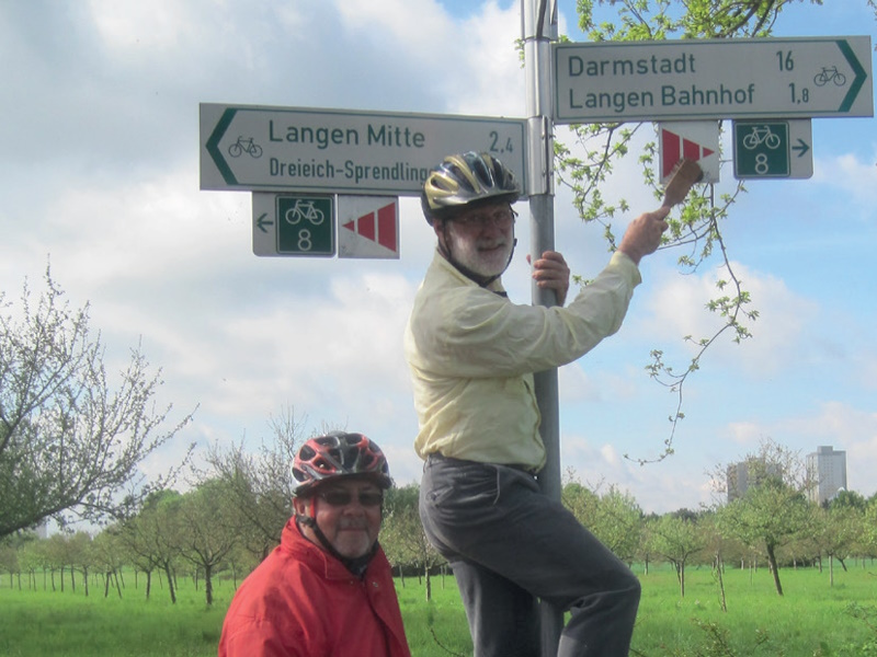 Zwei Personen mit Fahrradhelmen stehen an einem Radweg-Wegweiser mit Schildern nach „Langen Mitte“, „Darmstadt“ und „Langen Bahnhof“; ringsum eine grüne Streuobstwiese.