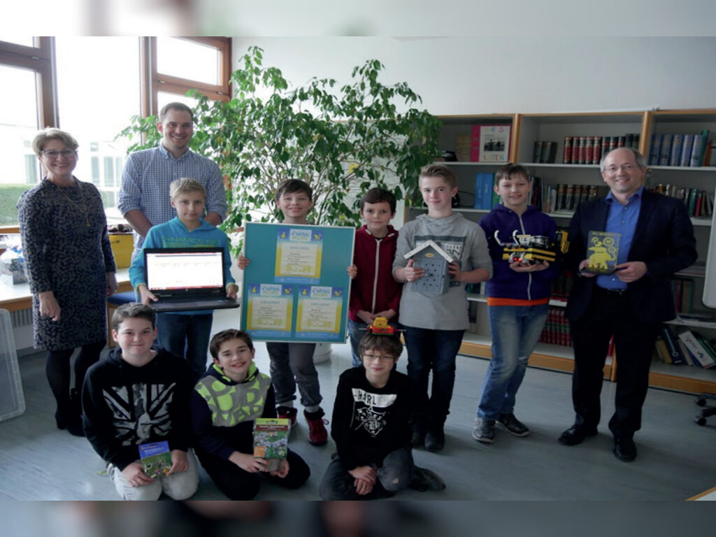 Eine Gruppe Kinder posiert mit drei Erwachsenen in der Schulbücherei; sie halten einen Laptop, eine Urkunde und selbst gebaute LEGO-Roboter in den Händen.