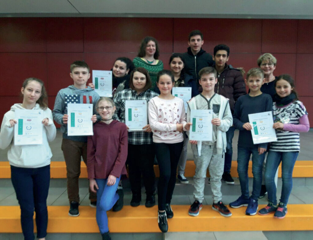 Eine Gruppe Kinder hält Urkunden in die Kamera, dahinter mehrere Lehrkräfte, auf den orangefarbenen Stufen im Schulforum.