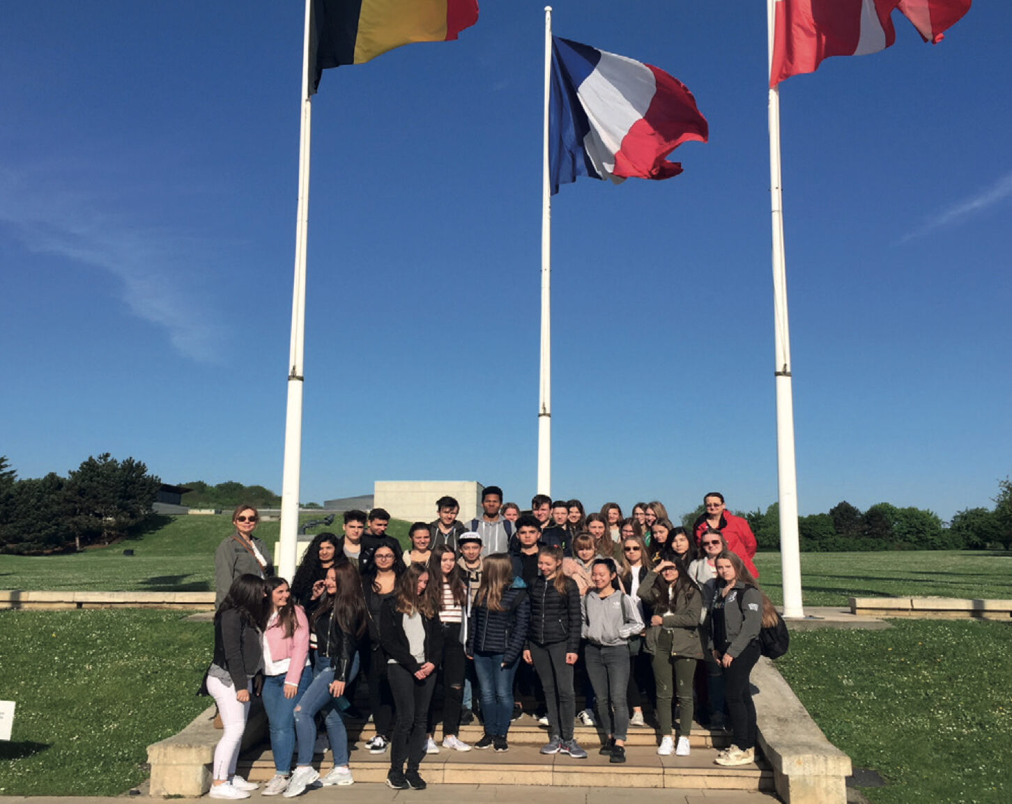 Eine große Schülergruppe posiert unter drei Fahnenmasten mit der belgischen, der französischen und einer weiteren Flagge auf einer Gedenkstätte mit weiten Rasenflächen.