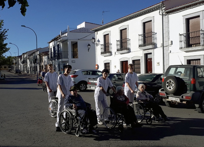 Mehrere junge Menschen in weißer Pflegekleidung schieben ältere Personen in Rollstühlen entlang einer sonnigen Straße mit weißen Häusern in einem spanischen Ort.