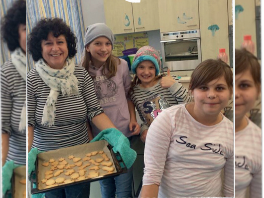 Eine erwachsene Person hält ein Blech mit frisch gebackenen Plätzchen; daneben stehen mehrere Kinder in einer Schulküche.