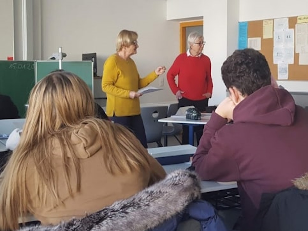 Zwei erwachsene Personen stehen vor einer Klasse und halten einen Vortrag; im Vordergrund hören Jugendliche an ihren Plätzen zu.