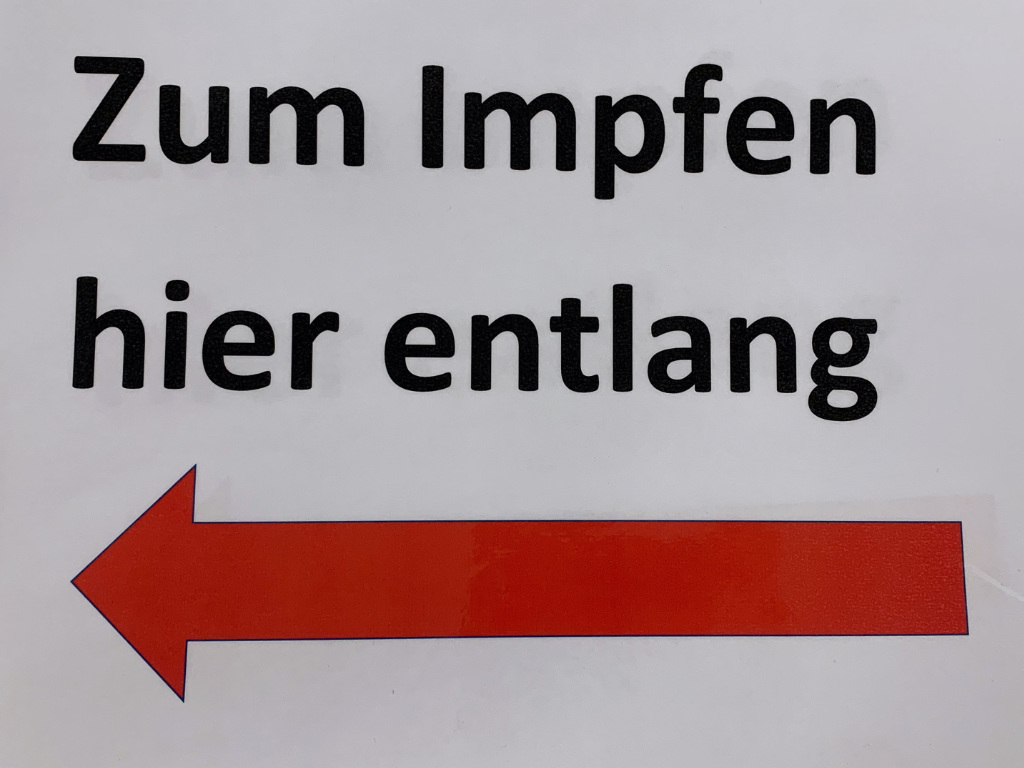 Ein Hinweisschild mit der Aufschrift „Zum Impfen hier entlang“ und einem großen roten Pfeil nach links.