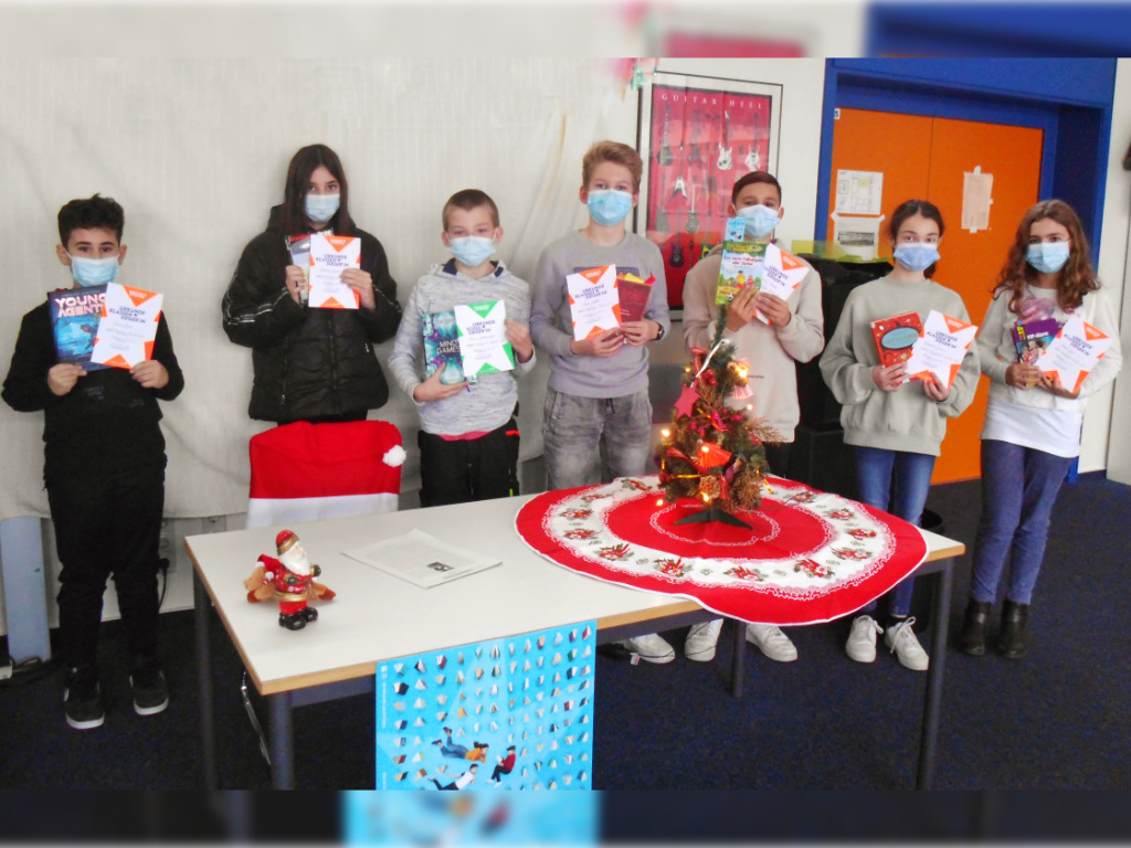 Sieben Kinder mit medizinischen Masken halten Urkunden und Bücher in die Kamera; auf dem Tisch davor steht ein kleiner geschmückter Weihnachtsbaum.
