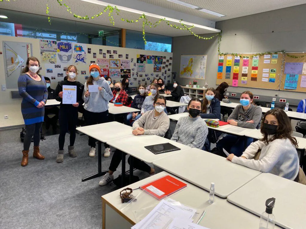 In einem Klassenraum hält eine Lehrkraft eine Urkunde hoch, die Klasse sitzt an den Tischen – alle tragen einen Mund-Nasen-Schutz.