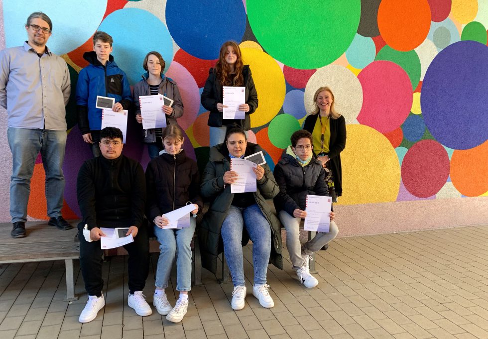 Eine Gruppe Jugendlicher hält Urkunden in die Kamera, an den Seiten zwei Lehrkräfte, vor einer bunt mit großen Kreisen bemalten Wand.