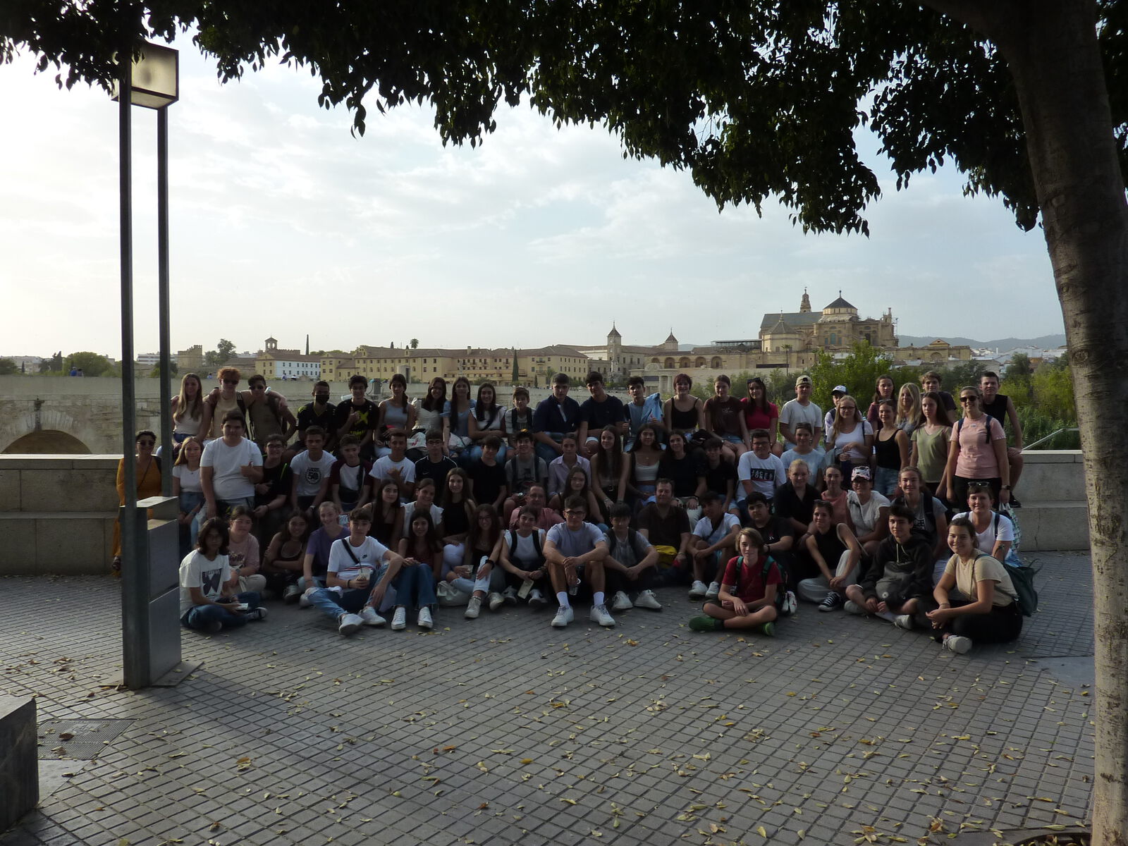 Eine große Schülergruppe sitzt und steht auf einem gepflasterten Platz; im Hintergrund die Mezquita-Catedral und die Römerbrücke von Córdoba.
