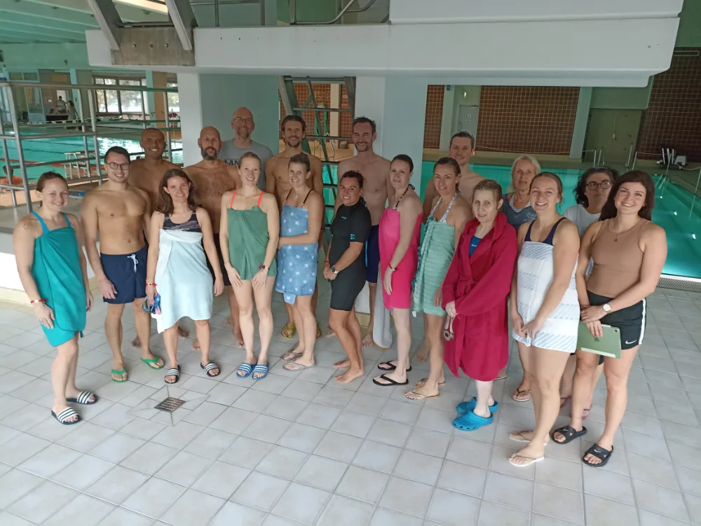 Die Teilnehmenden der Schwimmfortbildung stehen in Badekleidung und Handtüchern am Beckenrand des Hallenbads.