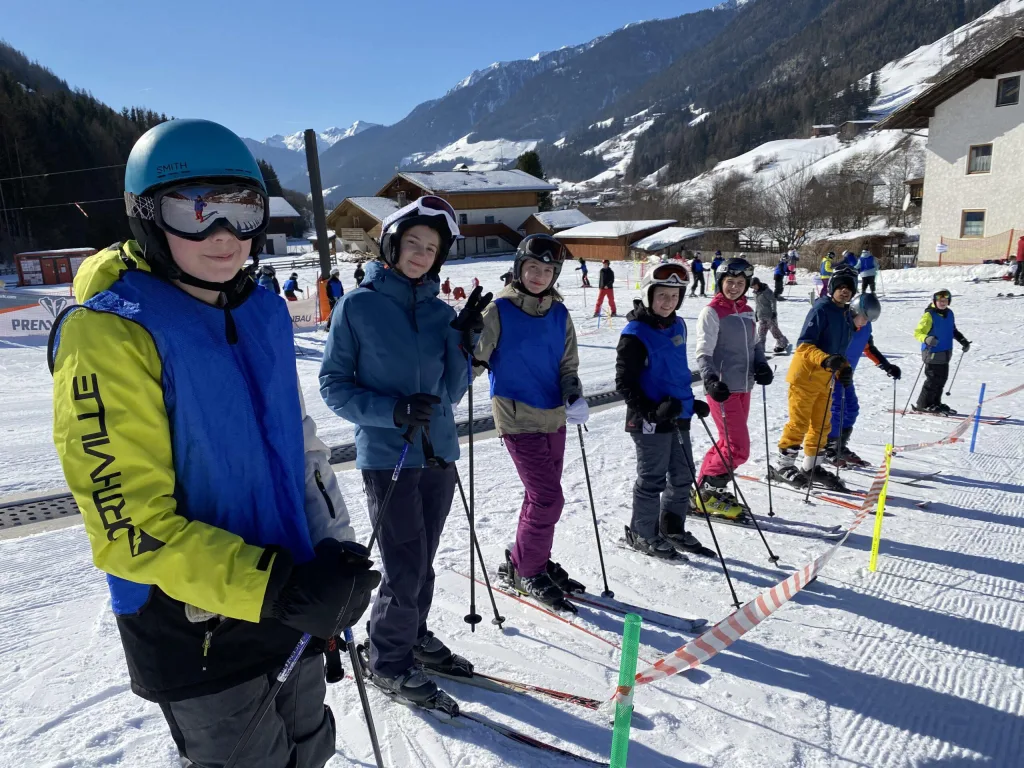 Jugendliche in Skikleidung mit Helm und Skibrille stehen mit ihren Skiern auf der Piste im Ahrntal, dahinter verschneite Berghänge.