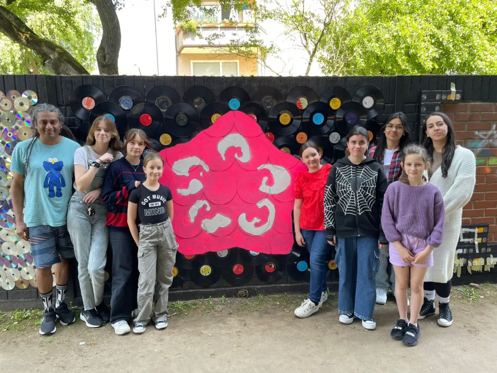 Die Kunstgruppe steht vor einer Wand aus alten Schallplatten, in deren Mitte eine große rosafarbene Blüte gestaltet ist.