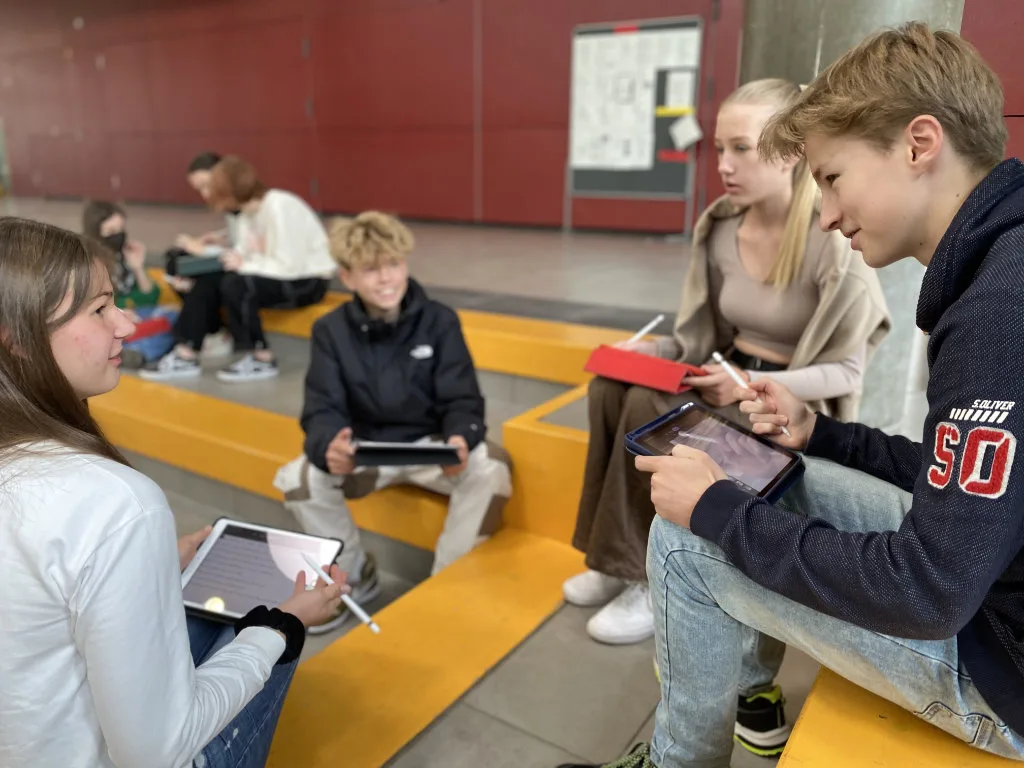 Jugendliche arbeiten mit Tablets auf den gelben Sitzstufen in der Pausenhalle.