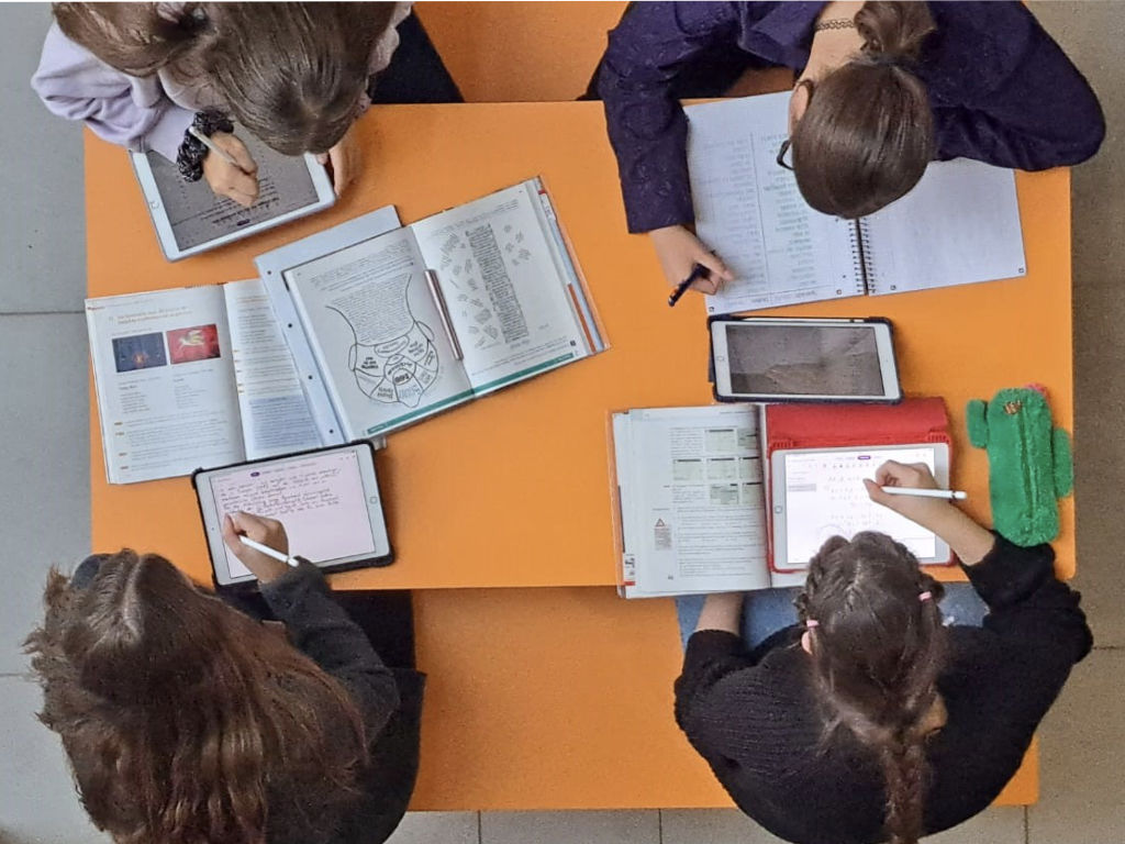 Blick von oben auf einen orangefarbenen Gruppentisch, an dem mehrere Jugendliche mit Tablets, Büchern und Heften zusammenarbeiten.