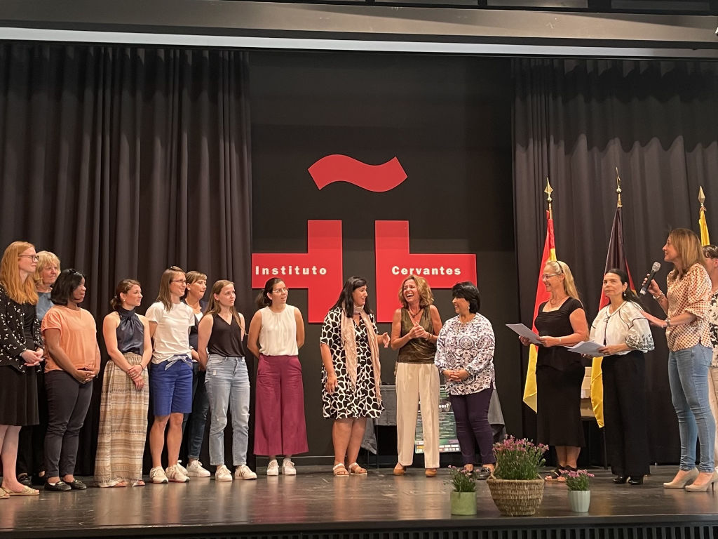 Eine Gruppe Erwachsener steht auf einer Bühne vor dem Logo des Instituto Cervantes und den Flaggen Spaniens und Deutschlands; eine Person spricht ins Mikrofon.