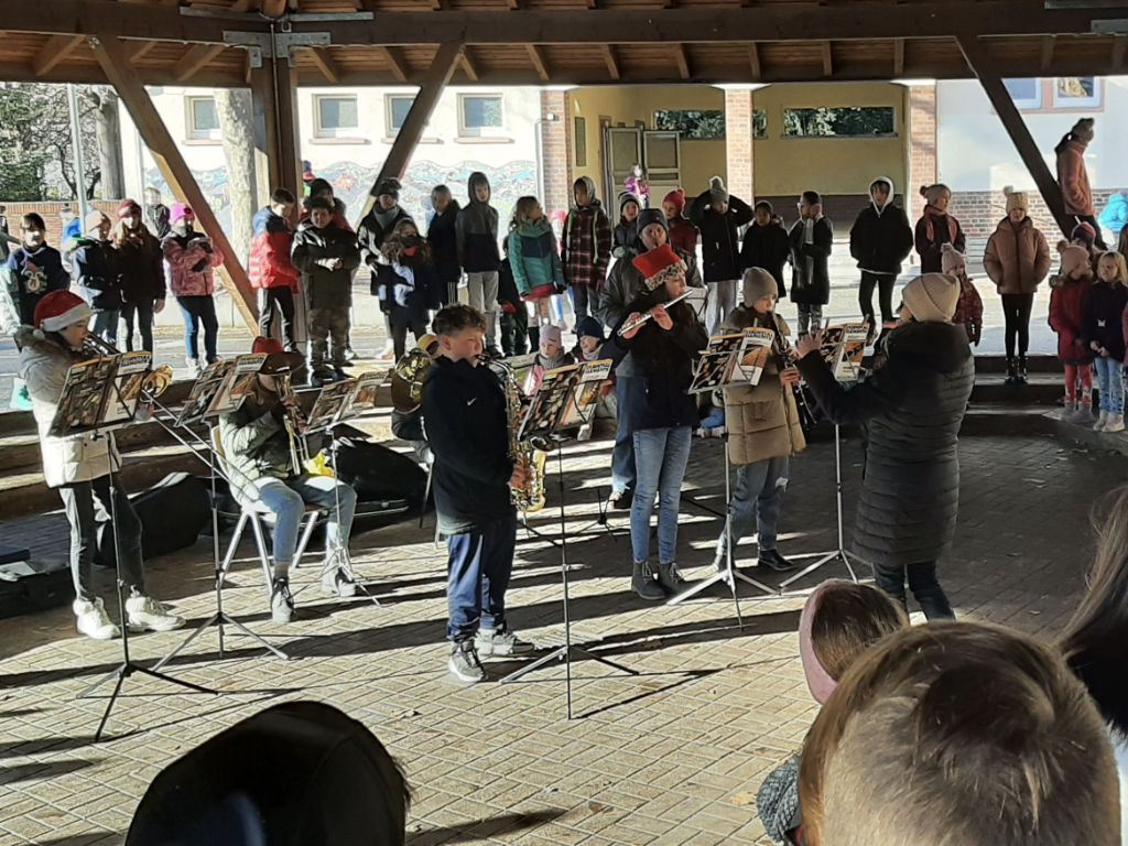 Eine Bläserklasse spielt unter dem überdachten Innenhof an Notenständern Blasinstrumente wie Saxophon und Trompeten; einige tragen Weihnachtsmützen, ringsum hört eine Gruppe Kinder zu.