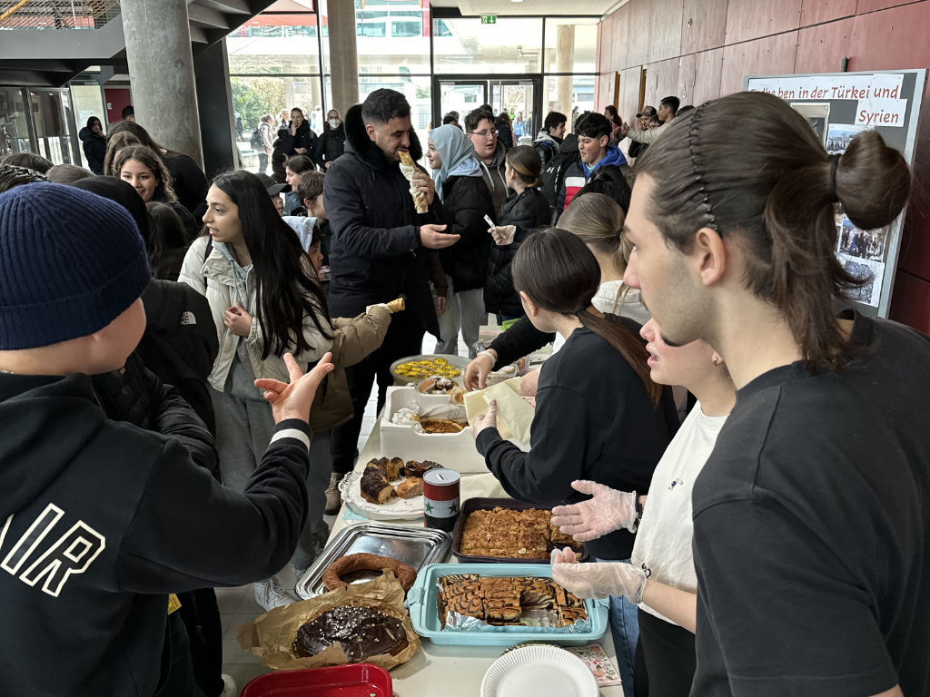 Reges Treiben an einem Kuchen-Verkaufstisch im Schulforum; Jugendliche verkaufen Gebäck, im Hintergrund ein Plakat mit der Aufschrift „Leben in der Türkei und Syrien“.