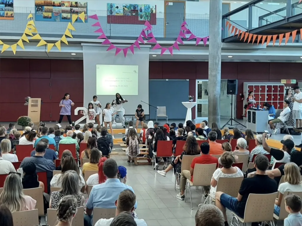 Die mit Wimpelketten geschmückte Aula ist bei der Einschulungsfeier bis auf den letzten Platz besetzt, vorne steht eine Gruppe auf der Bühne.