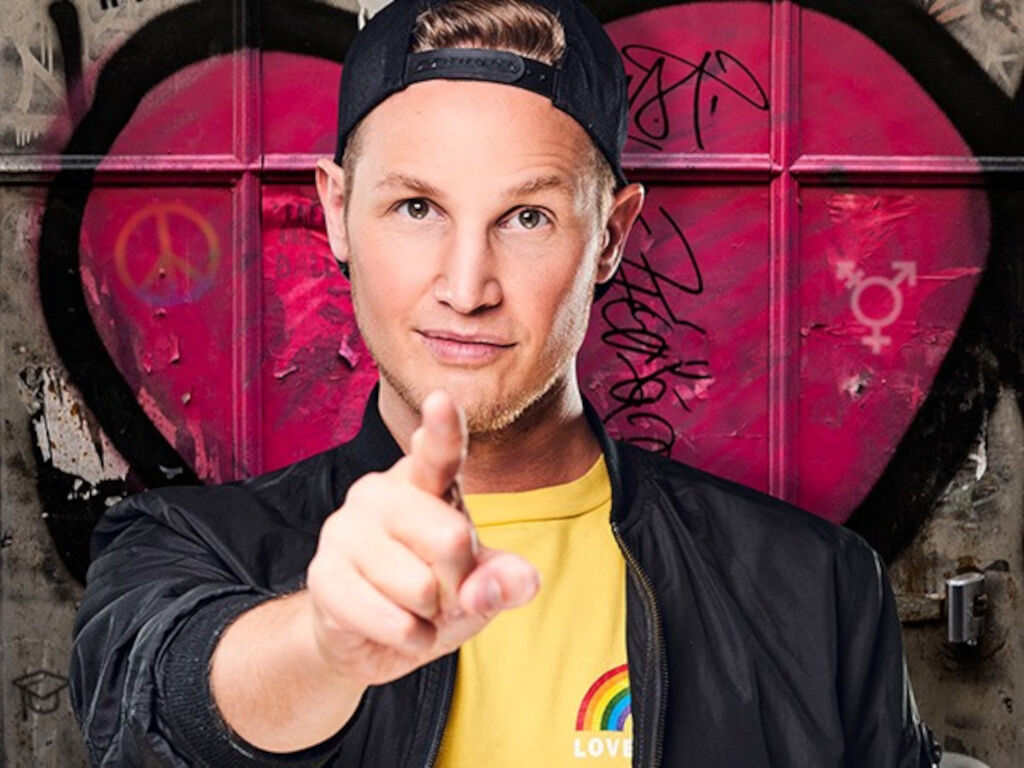 Ein Mann mit rückwärts aufgesetzter Cap und gelbem Shirt mit Regenbogen-Aufdruck zeigt mit dem Finger in die Kamera; im Hintergrund ein großes gemaltes Herz mit Friedens- und Transgender-Symbolen.