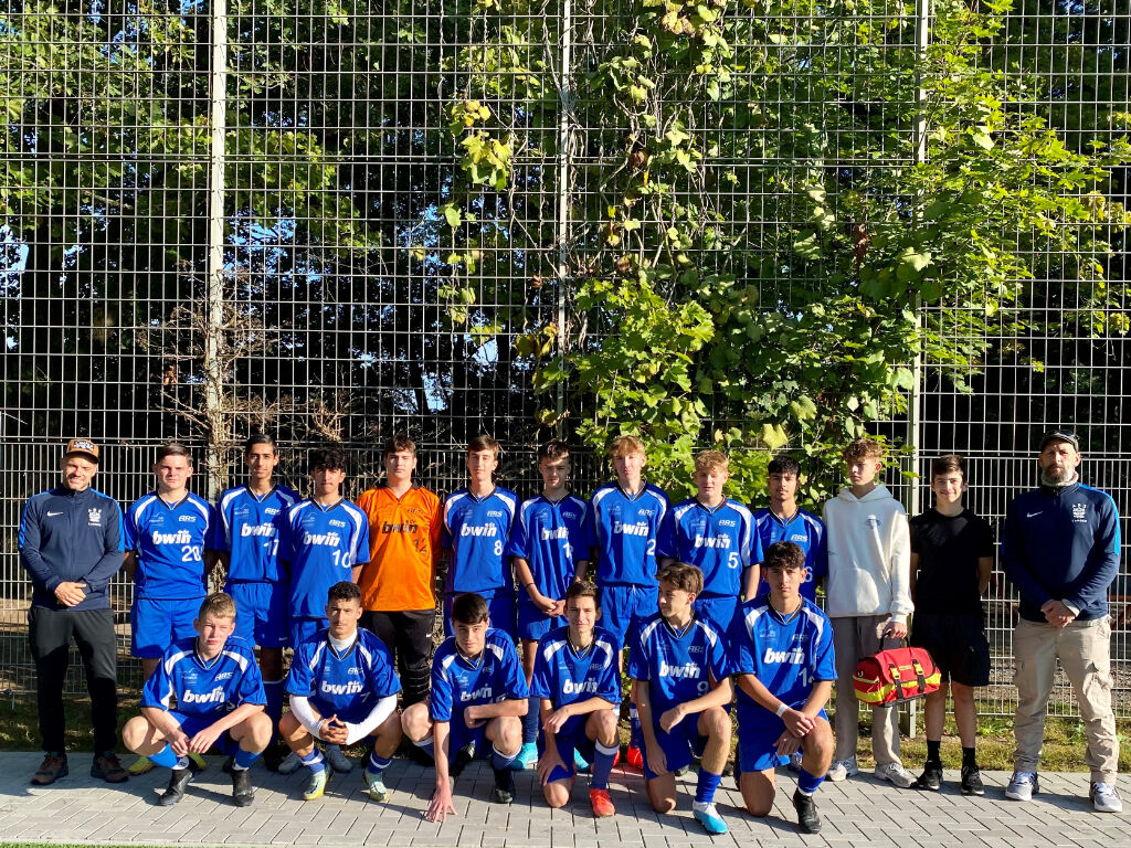 Die Schulfußballmannschaft in blauen Trikots mit dem Torwart in Orange posiert mit zwei Trainern vor einem Zaun; im Hintergrund Bäume.