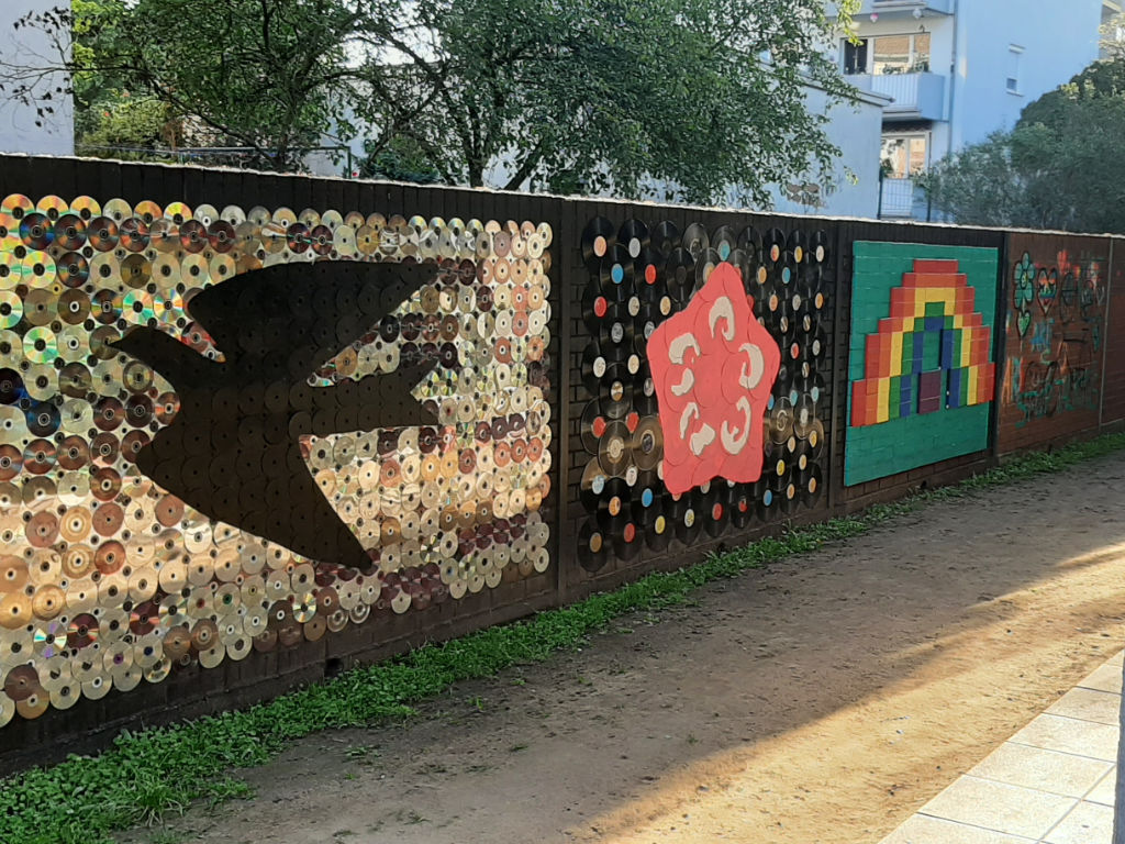 Eine lange Außenwand ist mit Mosaik-Kunstwerken aus glänzenden CDs gestaltet: unter anderem eine schwarze Vogel-Silhouette, ein rosa Stern und ein Regenbogen.