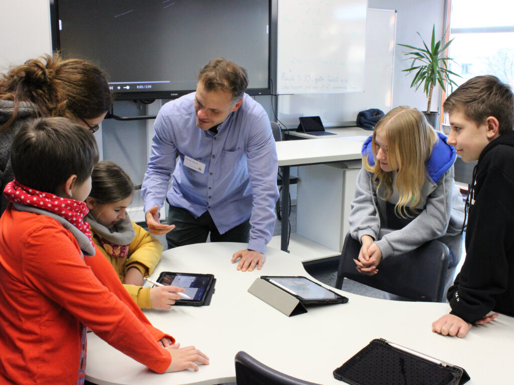 Eine Lehrkraft erklärt an einem runden Tisch mehreren Kindern und einer erwachsenen Person das Arbeiten mit iPads; im Hintergrund ein Smartboard.