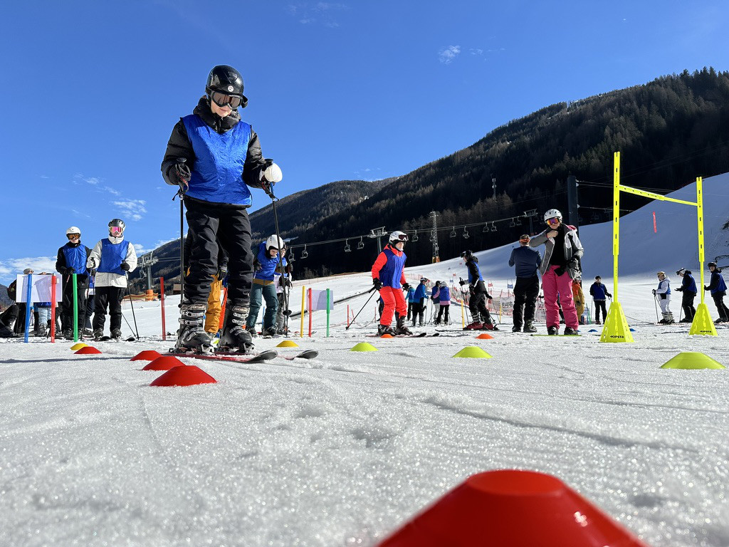 Eine Person auf Skiern fährt einen mit Pylonen abgesteckten Slalom-Parcours auf einer sonnigen Piste; im Hintergrund weitere Jugendliche und bewaldete Berge.