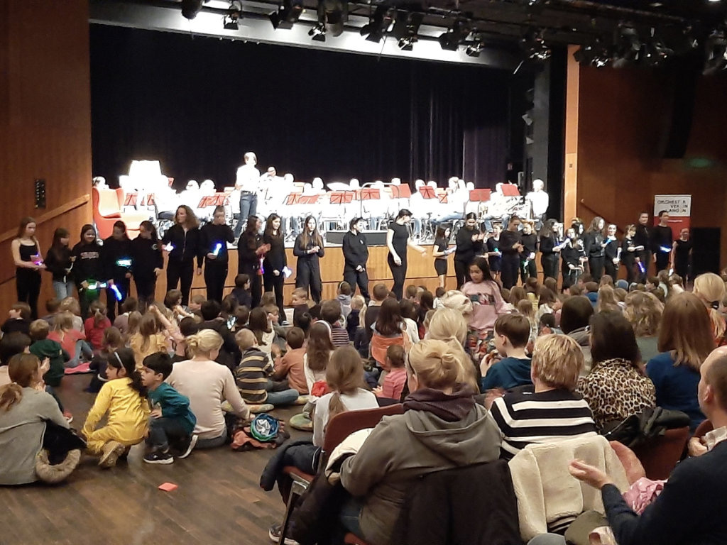Eine Aufführung in einem Saal: im Hintergrund spielt ein Orchester, davor steht eine Reihe schwarz gekleideter Mitwirkender mit leuchtenden Stäben; im Vordergrund sitzen viele Kinder als Publikum auf dem Boden.