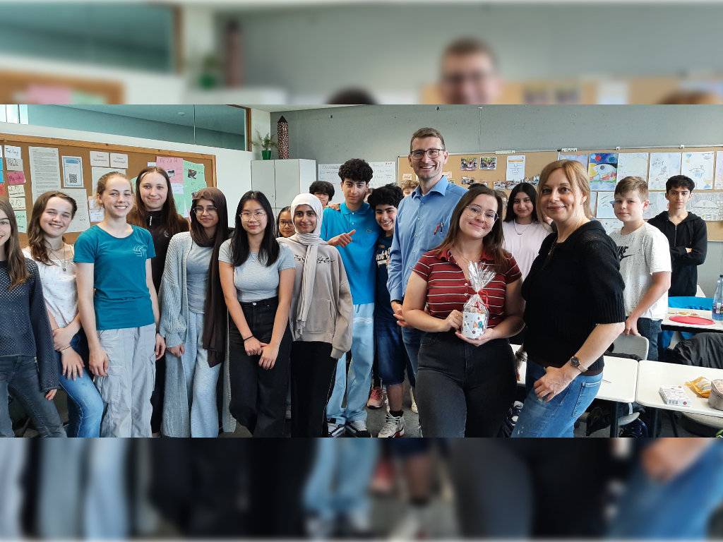 Eine Klasse posiert mit zwei Erwachsenen in einem Klassenraum; eine der erwachsenen Personen hält ein eingepacktes Abschiedsgeschenk in den Händen.