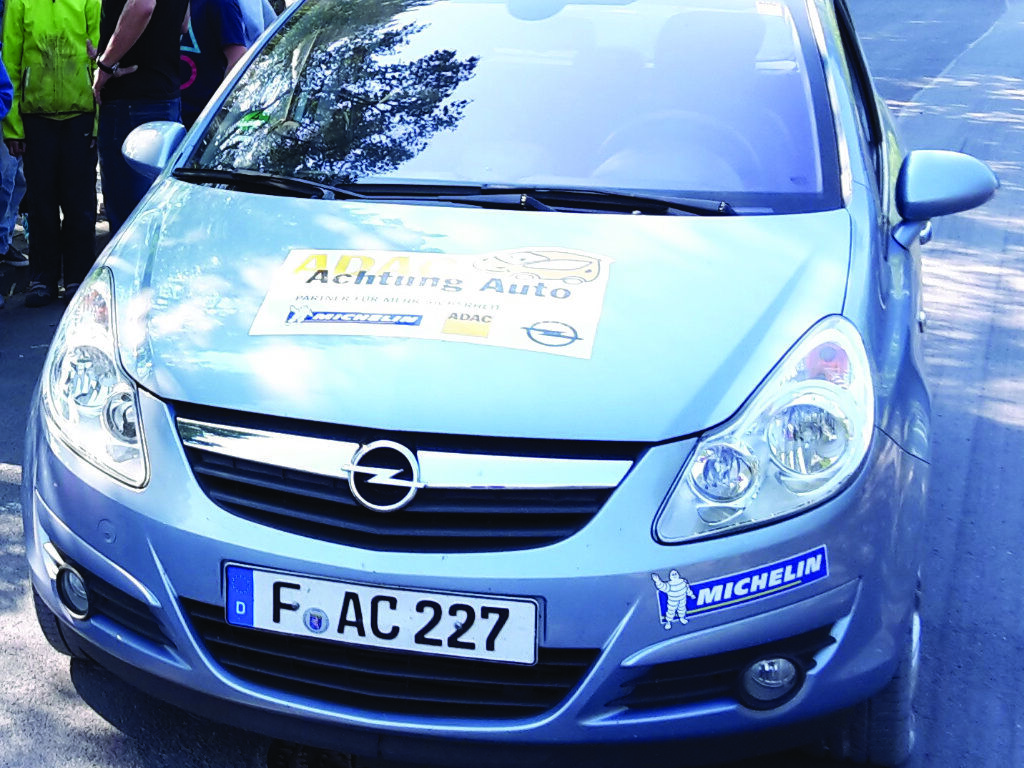 Ein silbernes Auto mit einem Aufkleber „ADAC Achtung Auto“ auf der Motorhaube; im Hintergrund stehen mehrere Kinder.