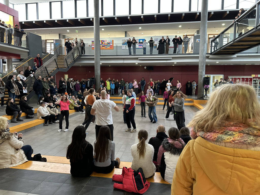 Im Schulforum führt eine Gruppe in der Mitte einen Tanz vor; ringsum sitzen und stehen zahlreiche Zuschauende auf den Stufen und der oberen Galerie.