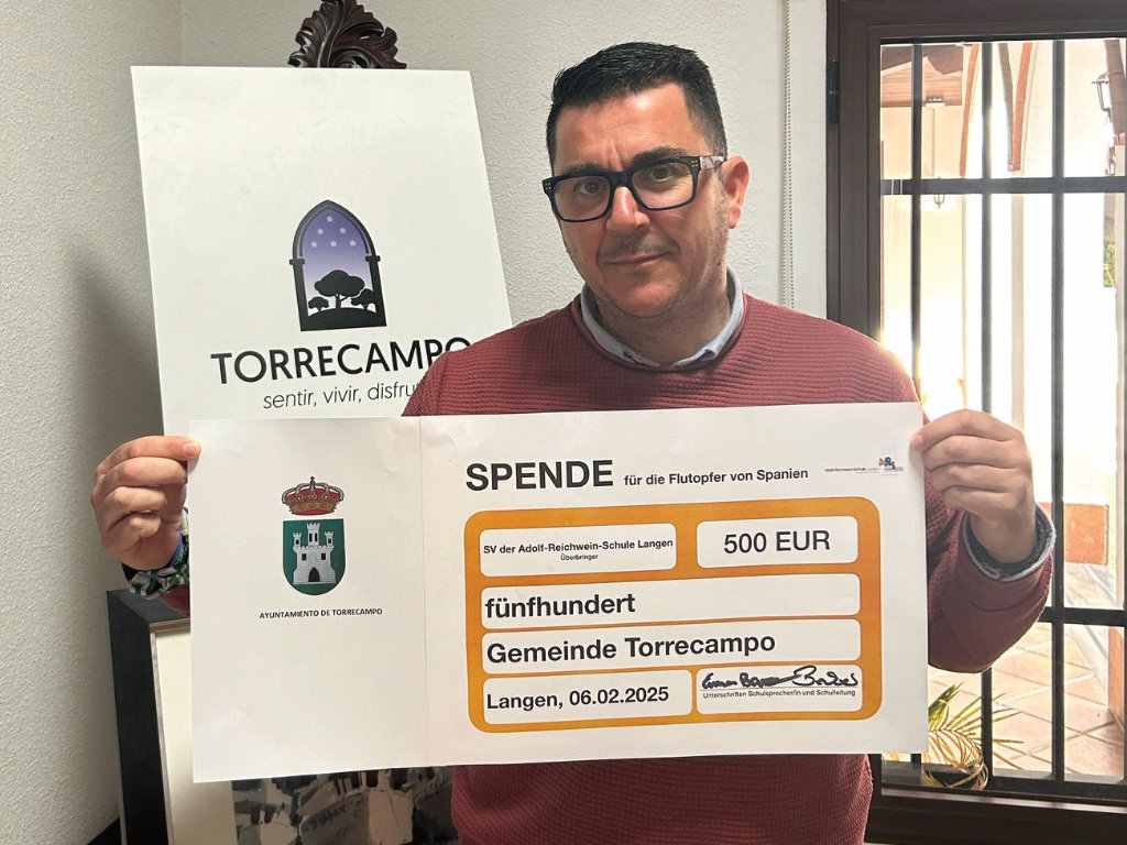 Eine Person hält einen großen symbolischen Spendenscheck über 500 Euro mit der Aufschrift „SPENDE für die Flutopfer von Spanien – Gemeinde Torrecampo“; im Hintergrund ein Plakat der Gemeinde Torrecampo.