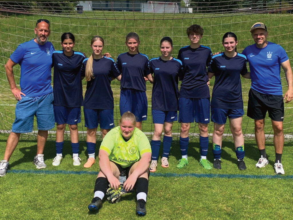 Die Mädchen-Fußballmannschaft in dunkelblauen Trikots steht mit zwei Trainern vor einem Tor auf dem Rasenplatz; vorne sitzt die Torhüterin in neongrünem Trikot.