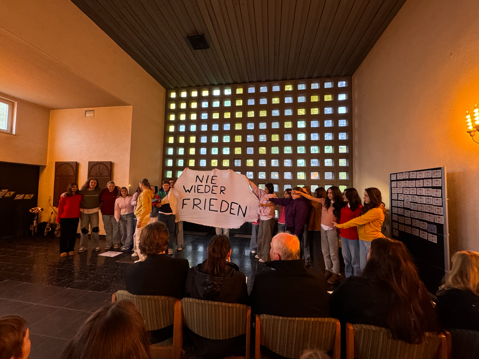 Eine Gruppe Jugendlicher steht in einem Saal und hält ein Tuch mit der Aufschrift „Nie wieder Frieden“ hoch; davor sitzt das Publikum, im Hintergrund eine Wand aus Glasbausteinen.